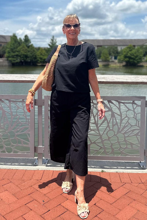 Woman wearing black loose fit co-ord set with wide-leg pants and short sleeves, standing outdoors by a railing on a sunny day