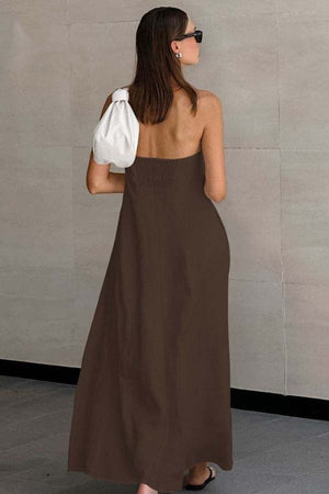 Woman wearing brown strapless long dress made from breathable cotton linen blend fabric, carrying white handbag, standing against gray tiled wall