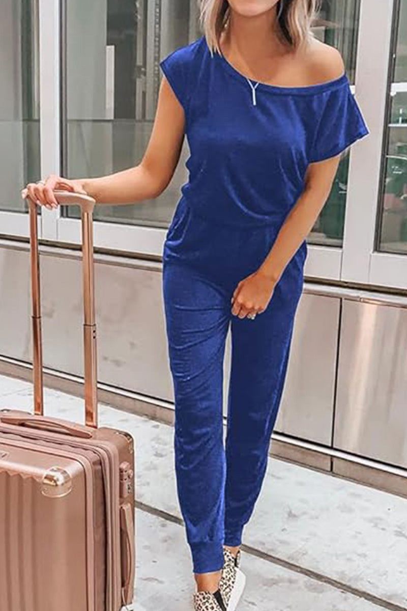 Woman wearing Moonlit Rome Off-Shoulder jumpsuit in blue, standing with suitcase indoors