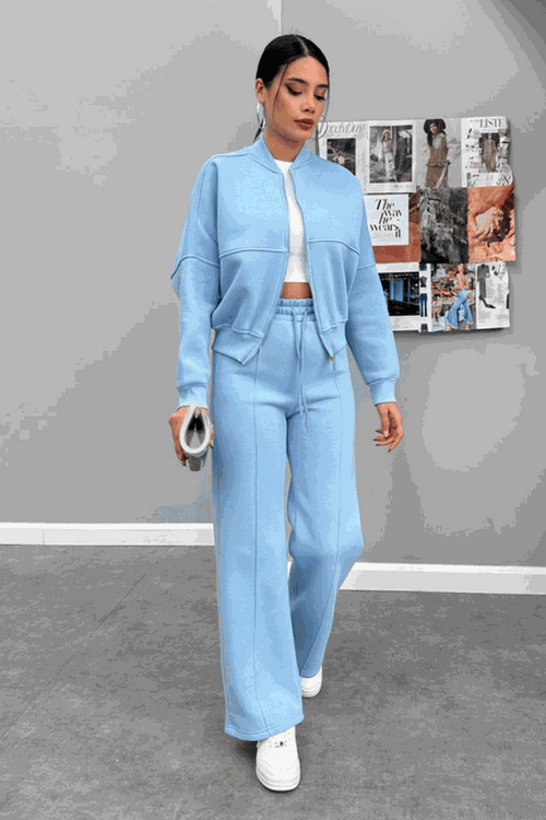 Woman wearing light blue The Voyager Zip Set with zip-front sweatshirt and wide-leg joggers, styled with white sneakers, standing indoors.
