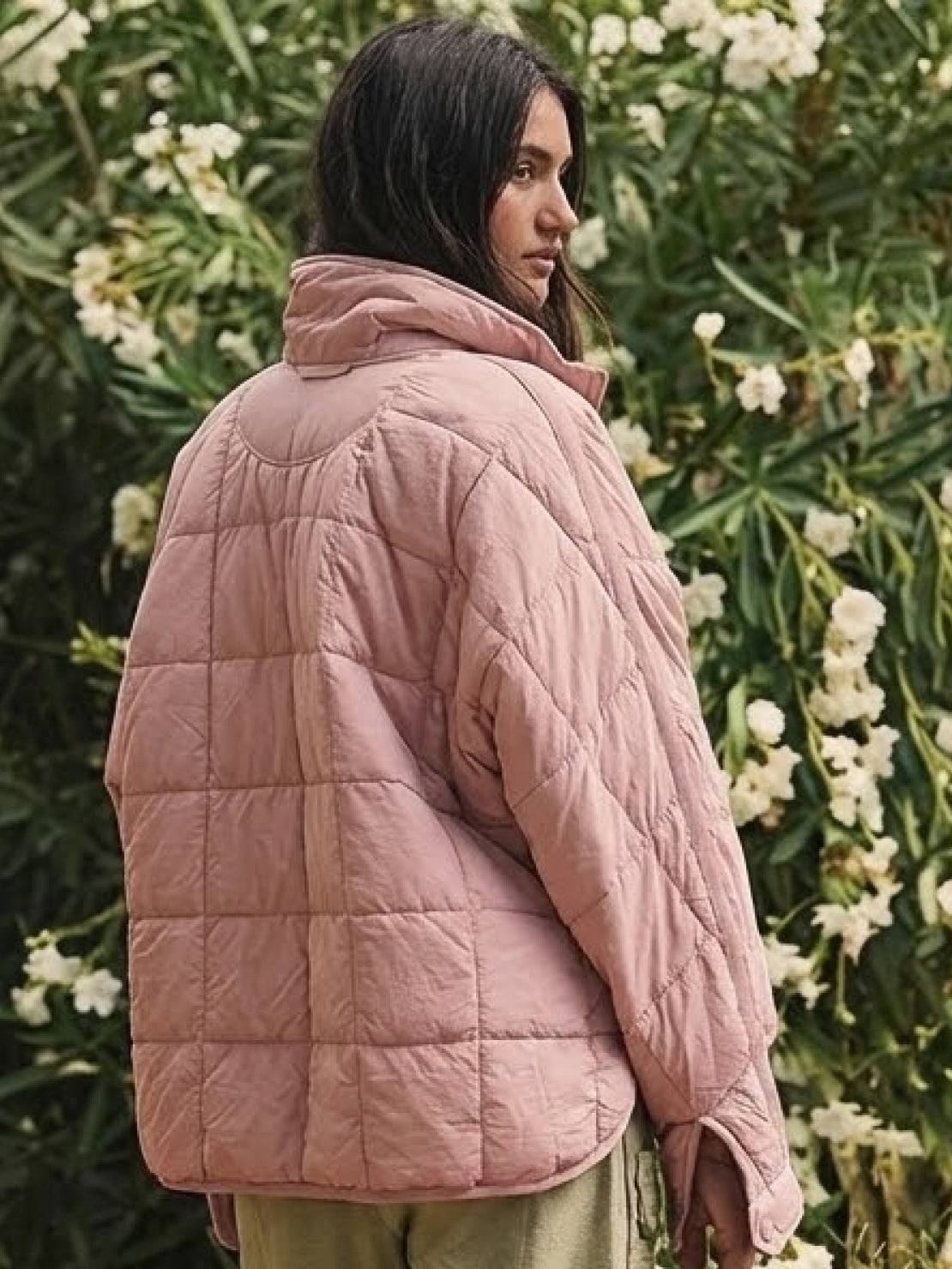 Woman wearing a blush pink lightweight puffer travel jacket standing outdoors near green foliage and white flowers.