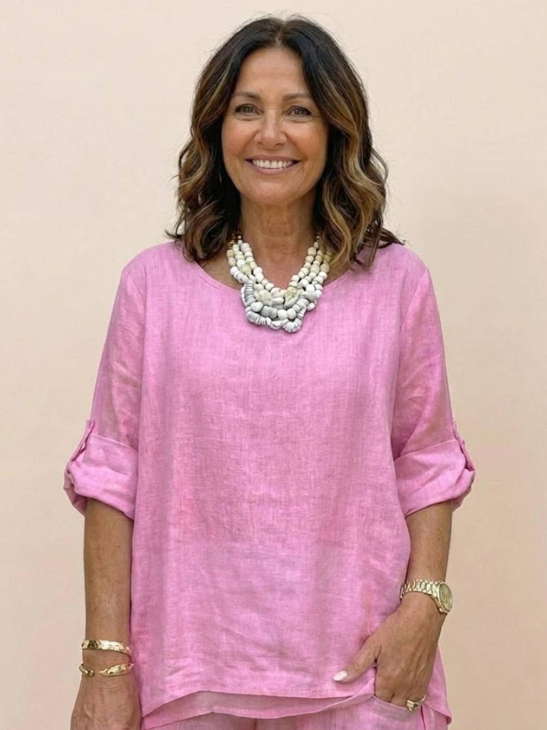 Woman wearing pink asymmetrical two-piece trouser set with rolled-up sleeves and statement necklace against plain background