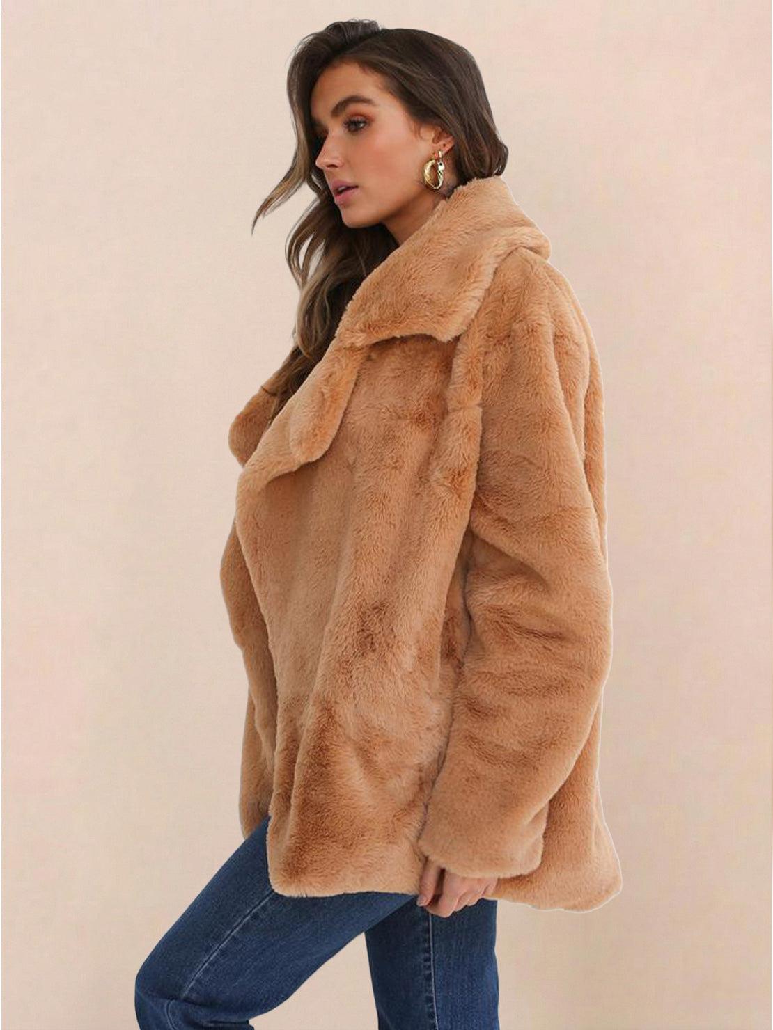 Woman wearing a tan open front oversized faux fur jacket with draped shawl collar and mid-hip length over blue jeans.