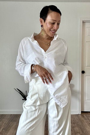 Woman wearing a white oversized linen shirt with relaxed fit and flowing hem paired with white pants indoors