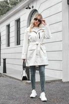 Woman wearing white water-resistant windbreaker trench jacket with belt, sunglasses, jeans, and white sneakers outdoors