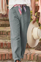 Woman wearing casual button-down utility-chic trousers with pockets and rolled cuffs, styled with a colorful scarf belt and holding a wide-brimmed hat.