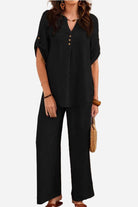 Woman wearing black wrinkle-free wide leg pants and button-front top set with short sleeves and gold buttons holding a woven handbag