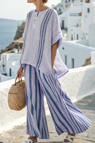 Woman wearing coastal comfort linen two-piece set with blue and white vertical stripes, loose fitting top and wide-leg pants, holding woven handbag outdoors