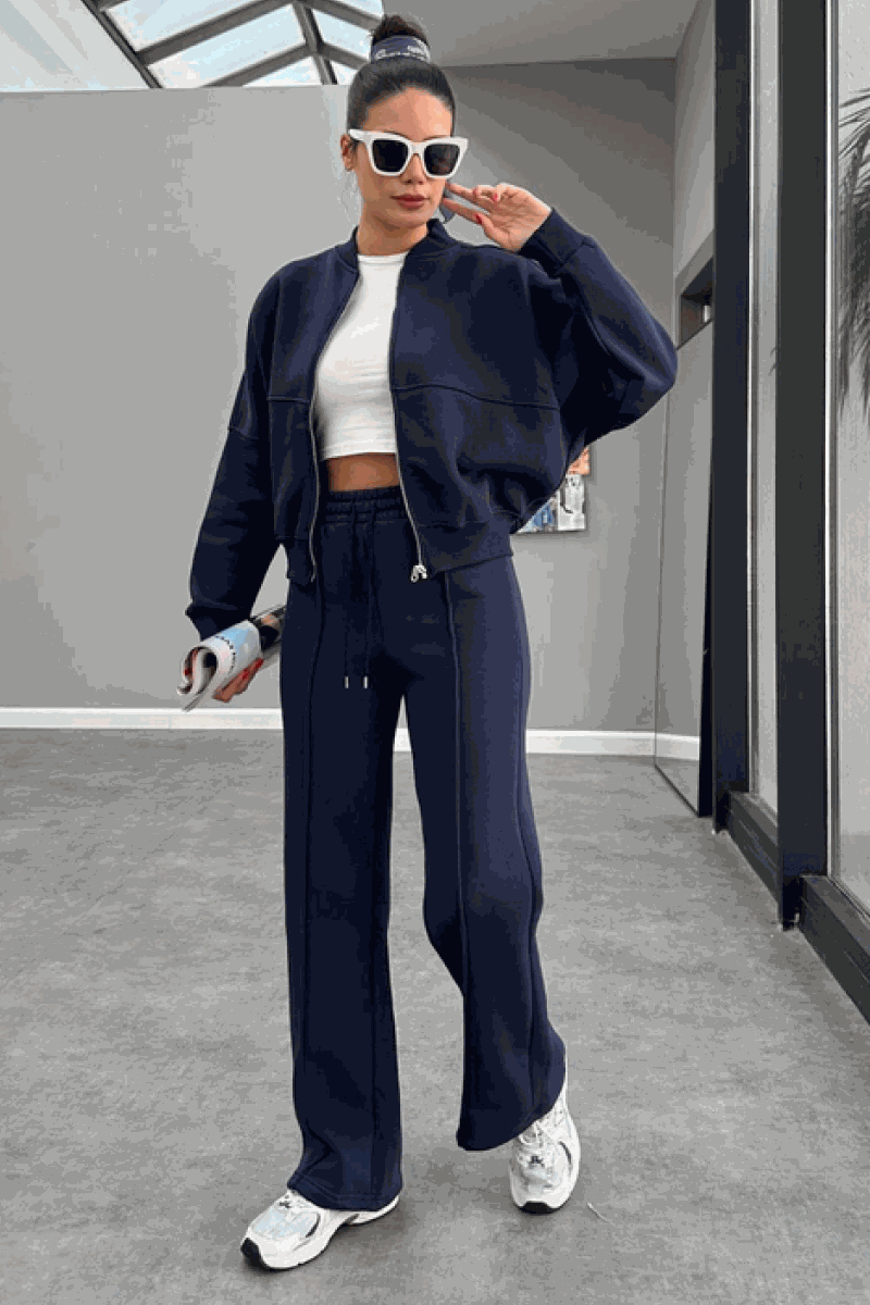Woman wearing The Voyager Zip Set navy zip-front sweatshirt and wide-leg joggers with sneakers and sunglasses in modern indoor setting
