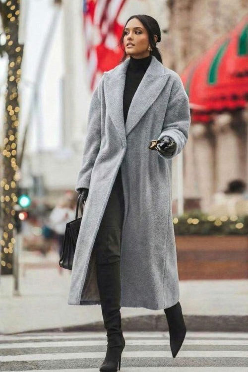 Woman wearing long grey oversized teddy coat with oversized lapels and black high-heeled boots walking on city street