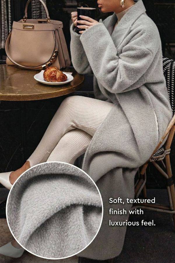 Woman wearing grey oversized teddy coat with soft textured fabric seated at café table with handbag and croissant