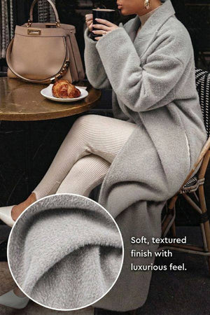Woman wearing grey oversized teddy coat with soft textured fabric seated at café table with handbag and croissant