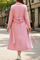 Back view of a pink timeless retro belted trench coat with a full-length cut and adjustable waist belt worn by a model walking on a city street
