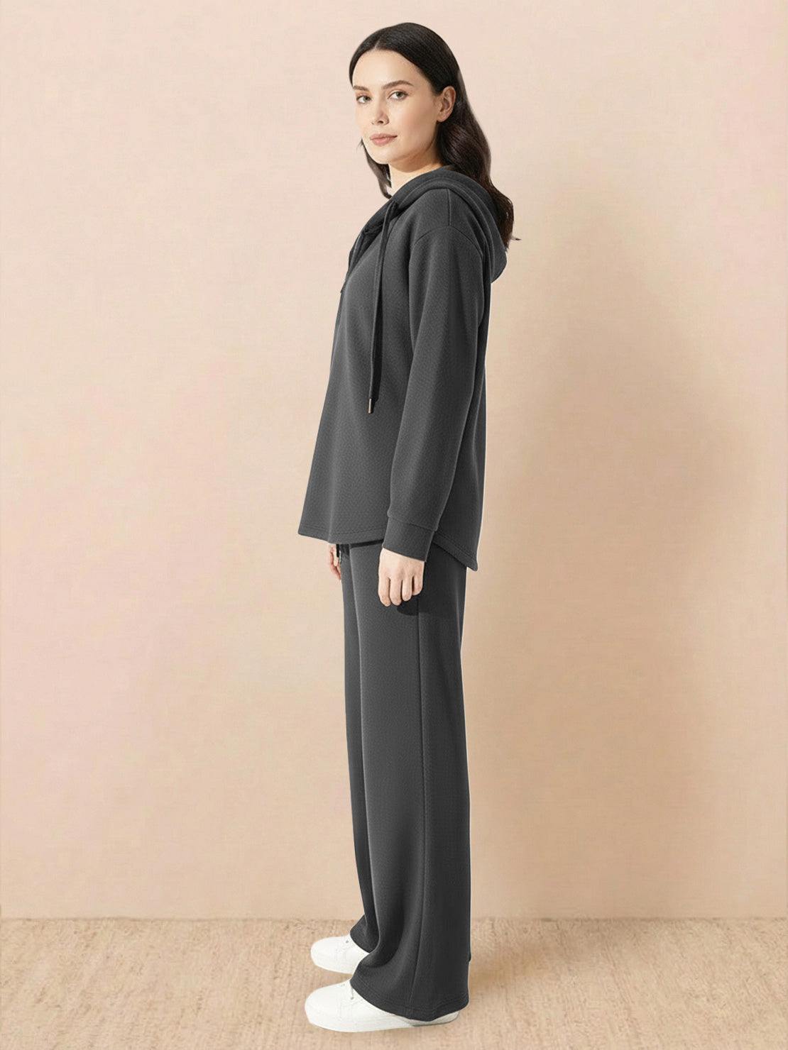 Woman wearing dark grey lyocell-blend hooded set with wide-leg pants and hoodie, standing sideways on beige background