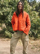 Woman wearing an orange lightweight puffer travel jacket outdoors with green foliage background