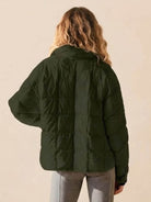 Back view of woman wearing dark green lightweight puffer travel jacket and gray pants against beige background
