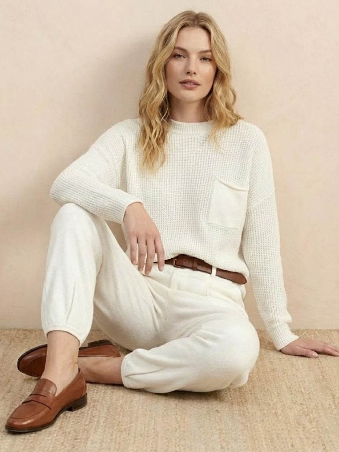 woman wearing European minimalism relaxed white knit sweater top and trouser set with brown belt and loafers sitting on floor