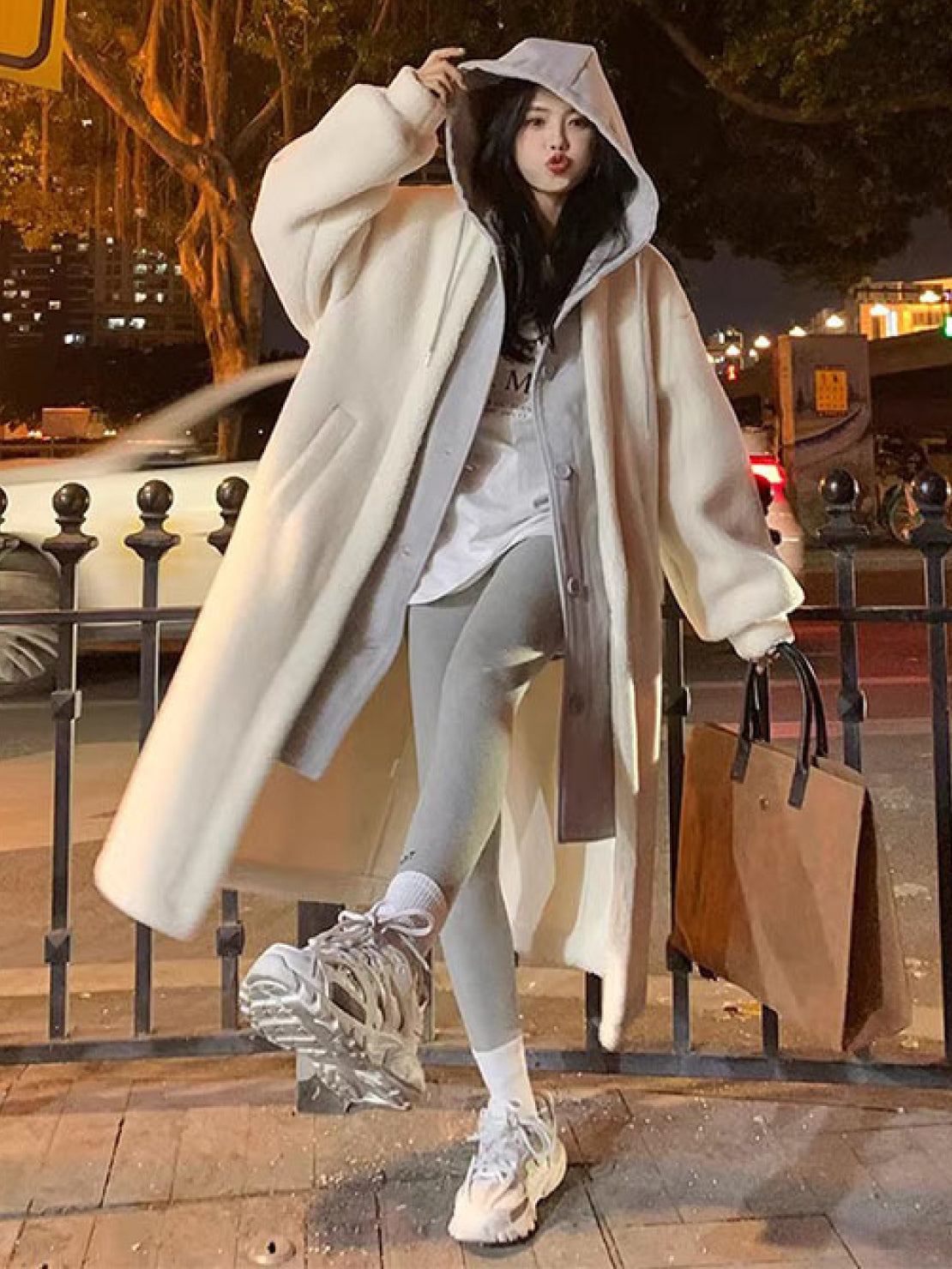 Woman wearing an oversized sherpa long coat with hoodie, layered over casual sporty outfit in urban night setting