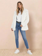 Woman wearing open front oversized white faux fur jacket with draped collar, paired with white top, blue jeans, and white sneakers against neutral background