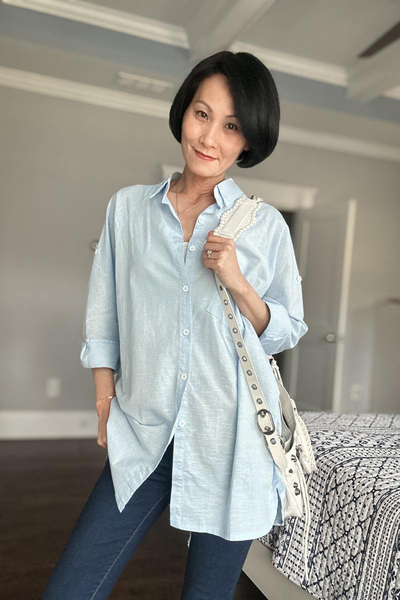 Woman wearing light blue oversized linen shirt with rolled sleeves and white shoulder bag indoors