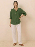 woman wearing green pure linen age-defying button detail shirt with white pants and sandals standing against beige background