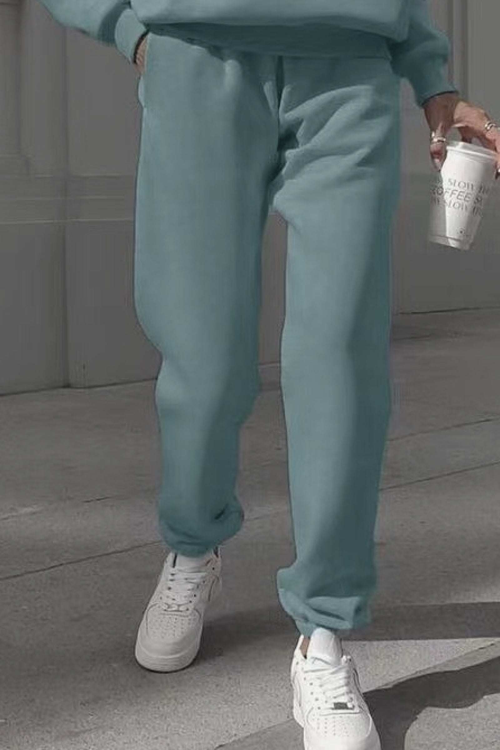 Woman wearing light green cotton blend tracksuit pants with white sneakers walking outdoors holding a coffee cup