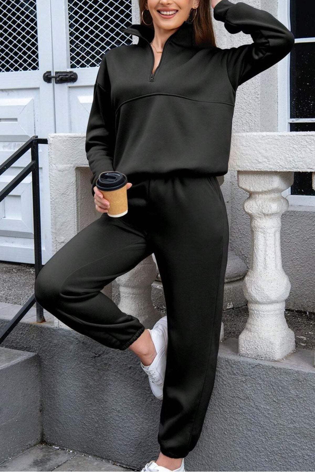 Woman wearing black cotton blend tracksuit with zip neckline and cuffed pants, holding coffee cup outdoors