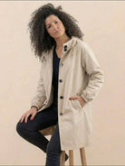 Woman wearing beige oversized Rain Couture Windbreaker water-resistant jacket with buttons and hood sitting on stool