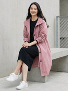 Woman wearing pink Rain Couture Windbreaker oversized water-resistant jacket with black dress and white sneakers sitting on concrete bench