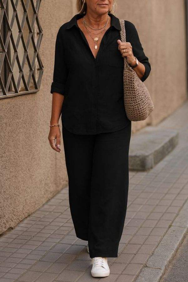 Woman wearing black relaxed shirt and trousers set with white sneakers and a woven tote bag walking outdoors