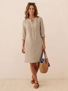 Women’s khaki cotton dress with 3/4 rolled sleeves and front buttons, styled with brown sandals and a woven handbag.