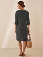 Back view of woman wearing knee-length charcoal cotton dress with rolled 3/4 sleeves, holding woven bag.