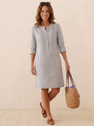 Woman wearing light gray cotton dress with 3/4 rolled sleeves and front pockets carrying straw tote bag